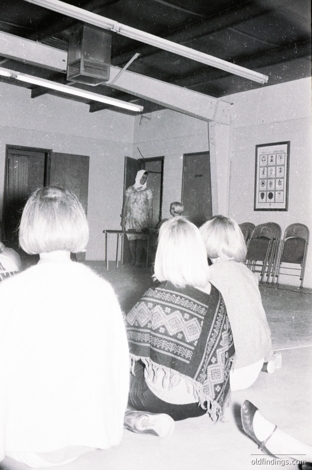 Mid-20th century classroom scene with teacher lecturing. Wooden benches, framed educational posters, and a projector mounted on ceiling. Audience faces teacher, one student wears patterned shawl. Institutional, formal atmosphere. Likely Eastern Bloc education system.
