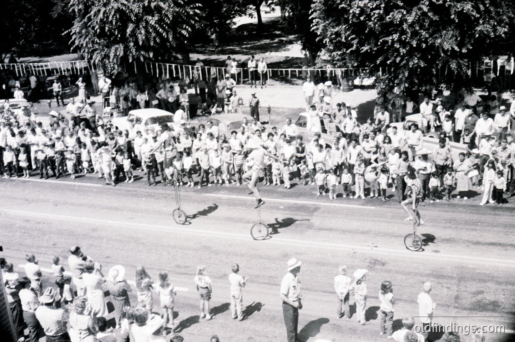 Aerial shot of a mid-20th-century unicycle performance in a park. Crowd of spectators lines fenced pathways, watching performers in motion. Lighting suggests daytime, likely 1950s–1970s.