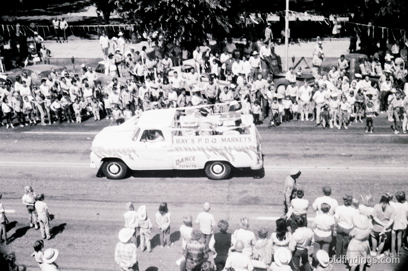 Vintage parade float featuring a retro station wagon branded with "Tray S.P.D.Q. Markets" and "Dance Tonight" in bold lettering, driving through a dense crowd. Mid-20th century street scene with spectators lining both sides, likely 1960s–1970s. Urban setting with visible trees and parked cars in background.