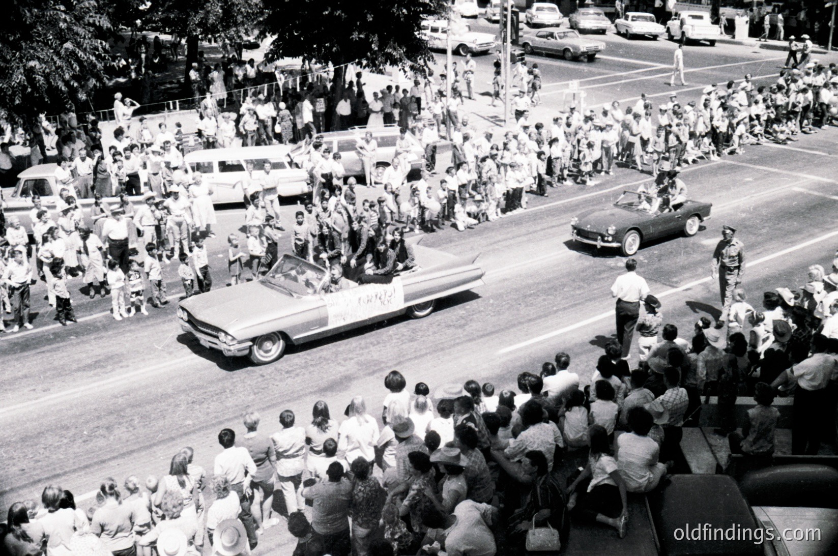 Vintage parade scene featuring a convertible car driving through a dense crowd, likely mid-20th century. Classic cars, spectators lining streets, and mid-block pedestrian barriers visible. Urban setting with trees and parked vehicles in background.