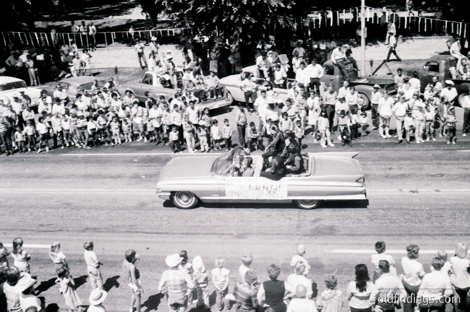 Classic 1960s convertible parade float with "Viva Kennedy" banner, surrounded by dense crowds. Mid-century American street scene with vintage cars and spectators in casual attire.