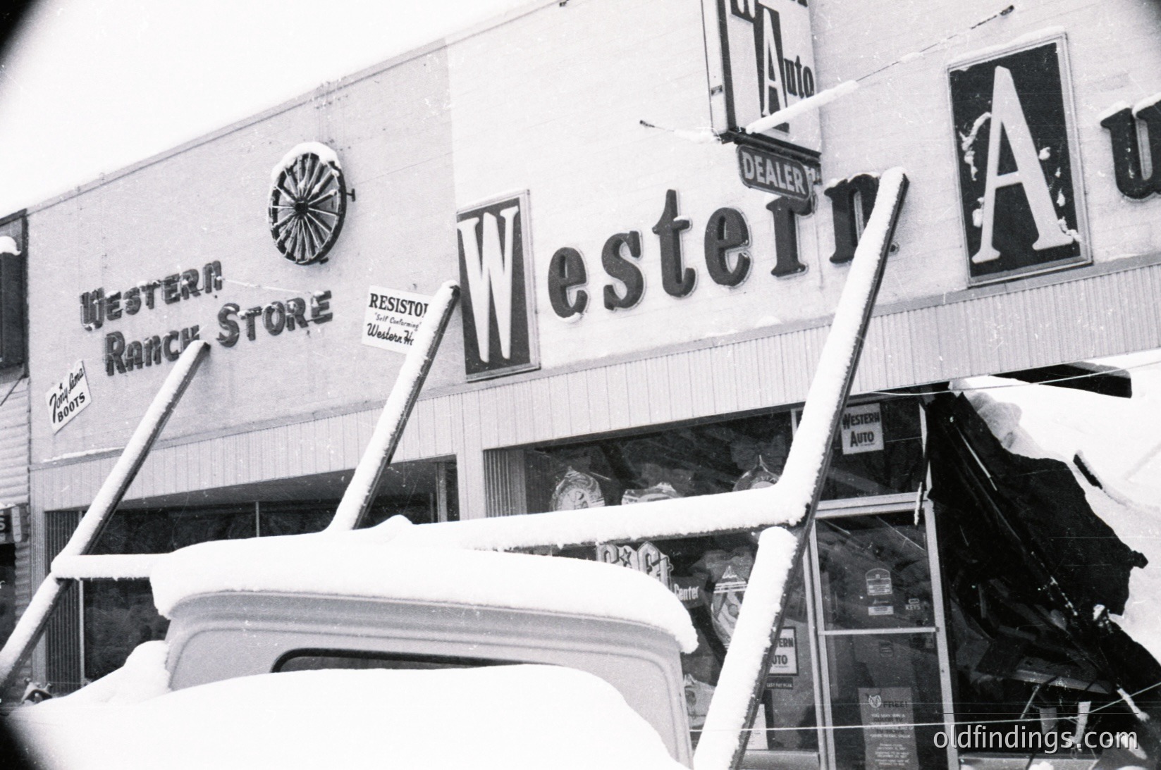 Vintage black-and-white photo of a Western Ranch Store sign with bold, retro lettering and a sunburst logo, dated mid-20th century. Snow-covered folding chairs and auto parts displayed outside. Resisto tires and auto dealer signage visible.