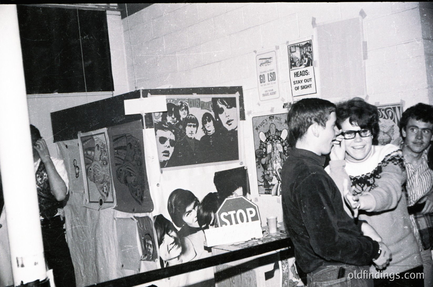 Indoor scene featuring three individuals in a dimly lit, cluttered room covered in posters and political art. Central figure wears glasses and a sweater, pointing at a "STOP" sign. Surrounding posters include anti-war and protest imagery, likely from the 1960s–1970s. One poster reads "Heads Stay Out of Vietnam."