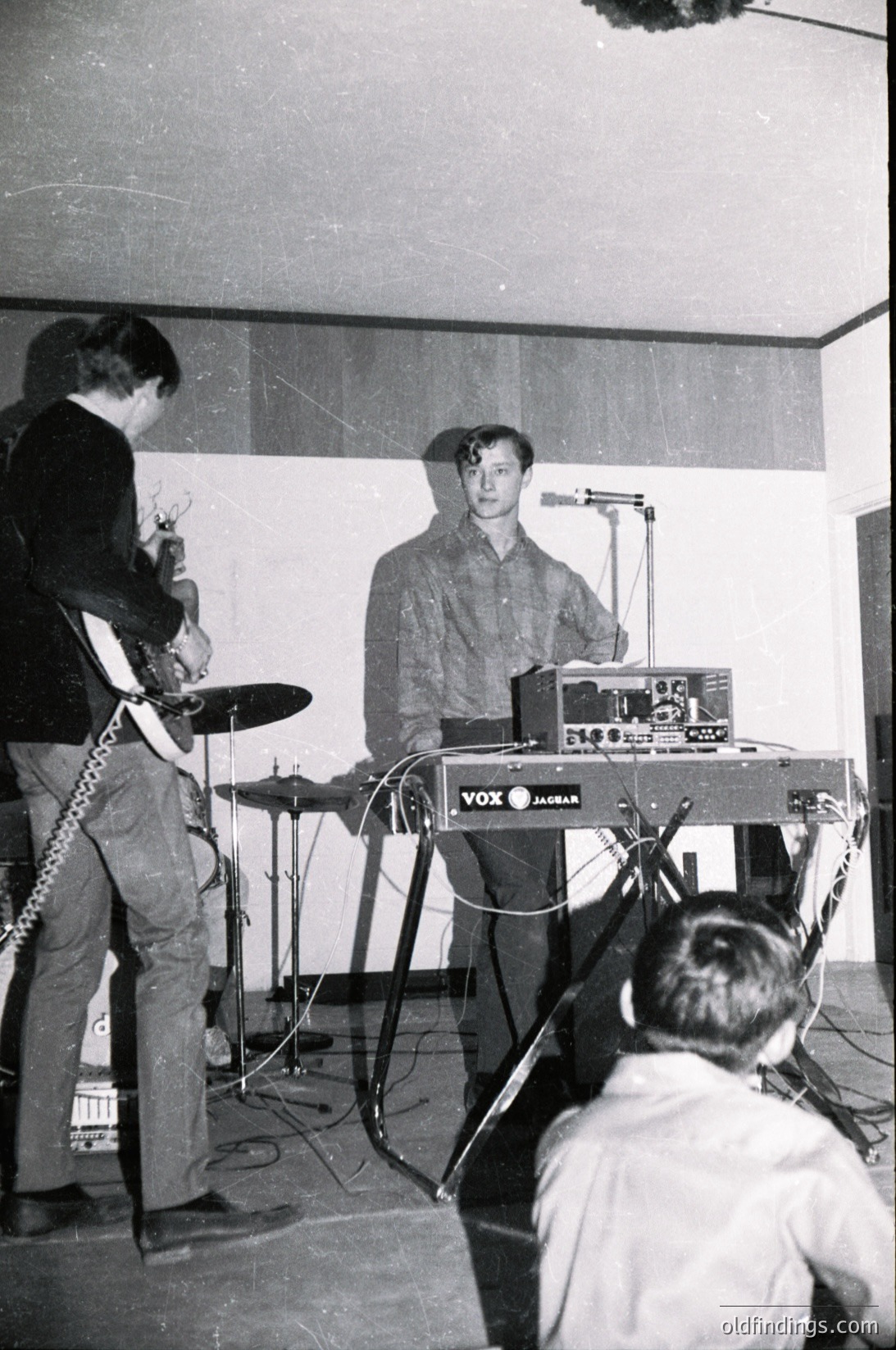 Vintage black-and-white photo of a live music performance featuring a guitarist (left) with a Fender-style bass, a keyboardist (center) playing a VOX Jaguar organ, and an audience member (right) facing the stage. Minimalist stage setup with basic lighting. Likely 1960s–1970s rock scene.
