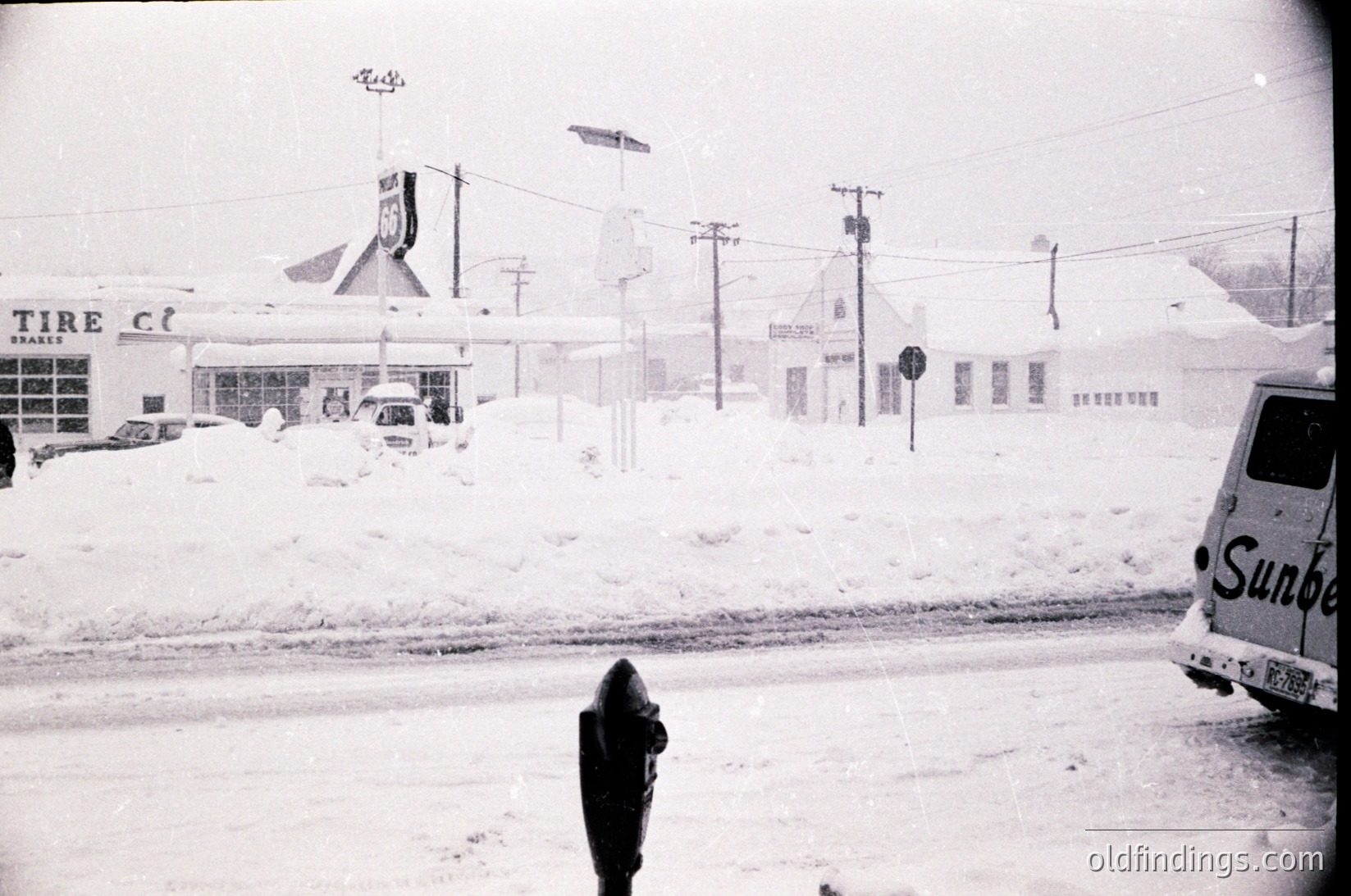 Mid-20th century snowstorm scene: blanketed gas station ("Tire Club") and adjacent buildings under heavy snowfall. Classic 1950s-60s American roadside architecture with vintage signage. Snow-covered "Sunoco" truck parked near a partially cleared road.