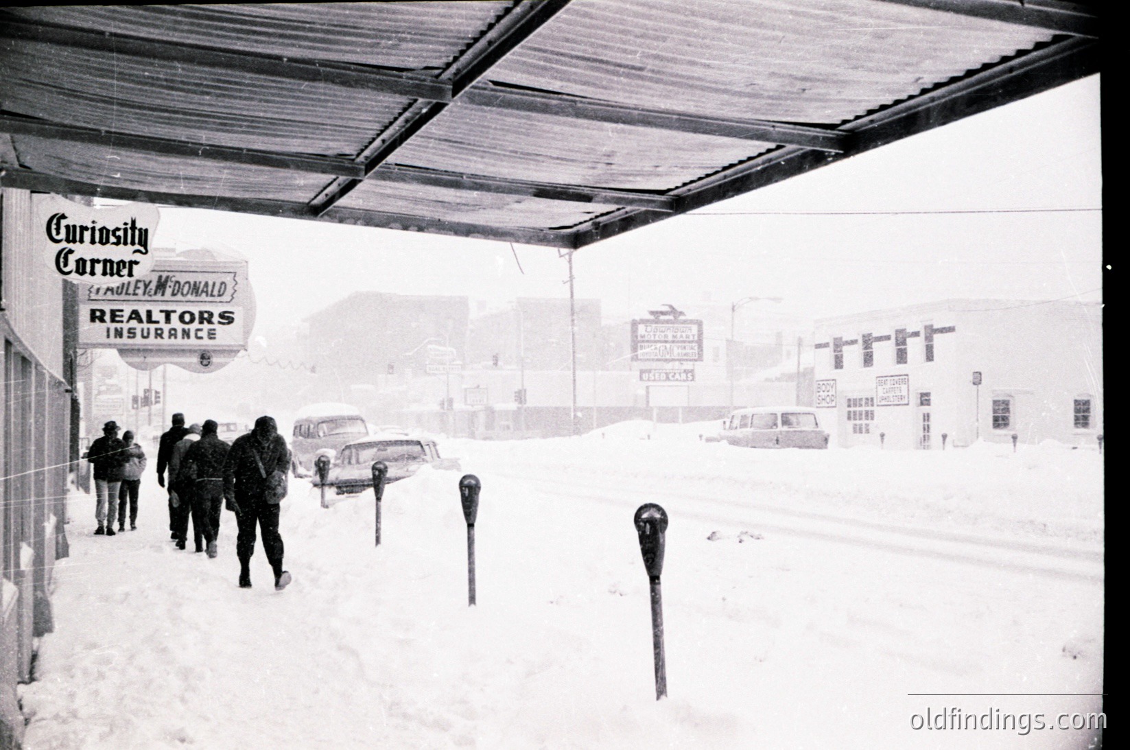Blizzard conditions at a mid-20th century highway rest stop, featuring a "Curiosity Corner" McDonald’s Realtors Insurance sign. Snow blankets parked cars, parking meters, and a covered walkway. Distant directional signs indicate routes 6 and 10. Classic vintage American roadside scene.