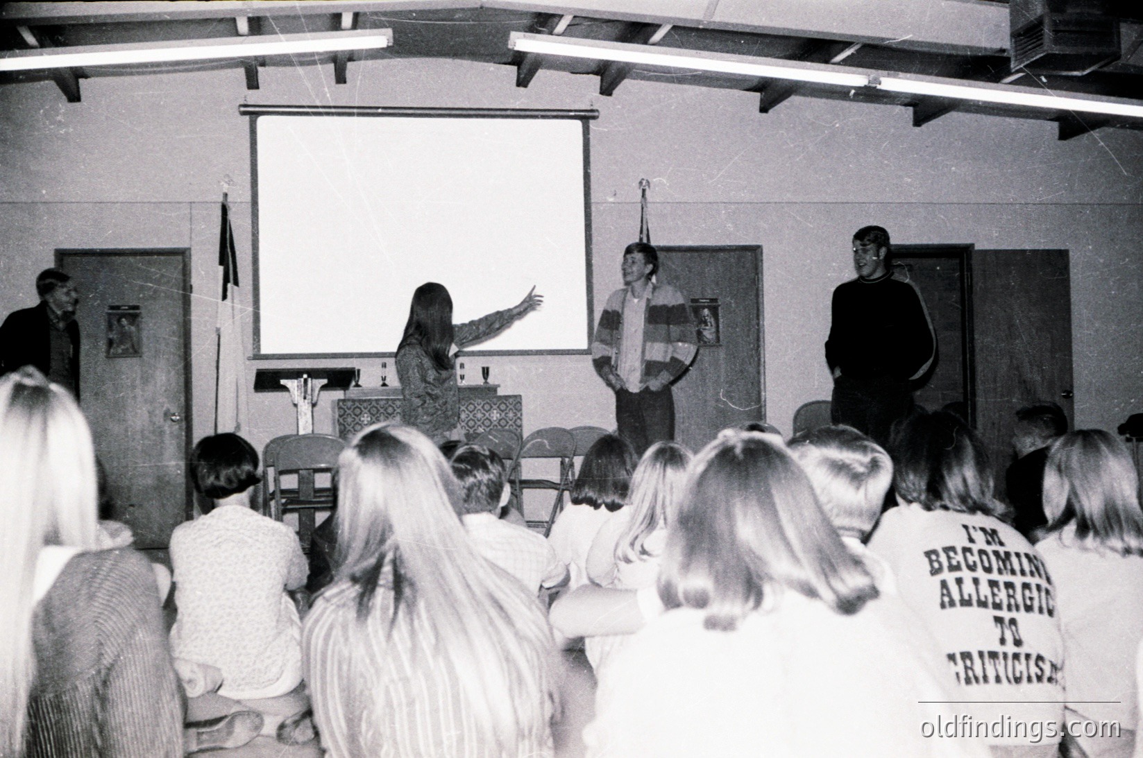 Black-and-white classroom scene featuring three lecturers at a projector screen, addressing seated students. Audience wears 1970s-style long-sleeve shirts; one visible tee reads *"I'm becoming allergic to criticism."* Wooden desks and simple stage setup suggest a university or workshop setting.