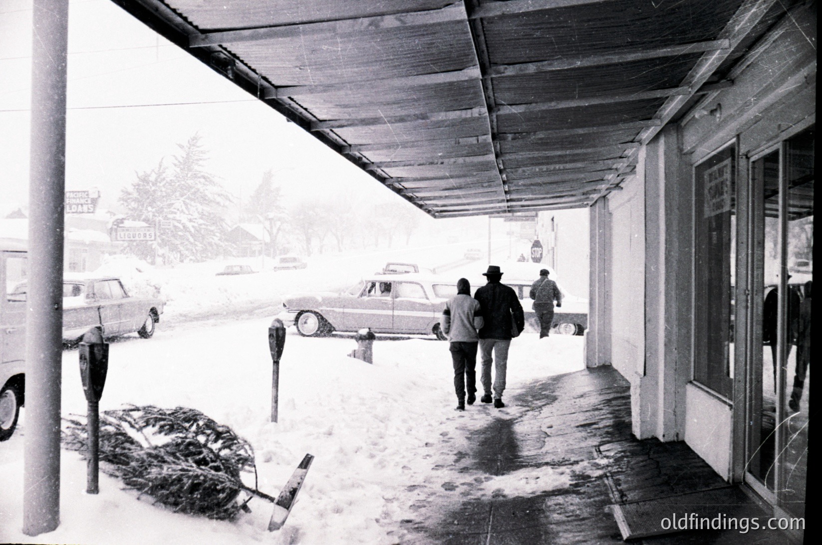 Mid-20th century street scene under a covered sidewalk during heavy snowfall. Three pedestrians in winter coats and hats navigate snow-covered pavement beside parked vintage cars (, , ). Snowbanks and a bundled firewood stack highlight winter preparedness. Commercial buildings with simple signage suggest a small-town atmosphere (, ).