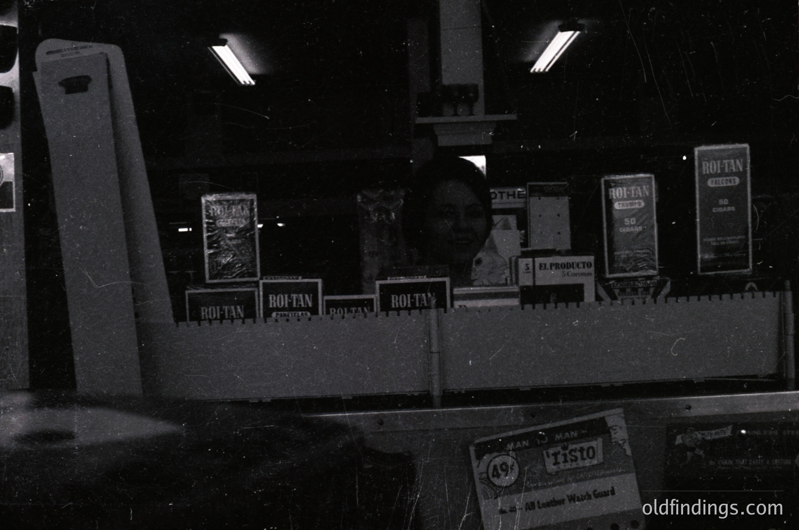 Vintage black-and-white photo of a dimly lit industrial or warehouse setting, featuring stacked "Rohtan" branded leather products (likely belts and saddles) on metal shelving. Visible signage includes "El Producto" and "TISTO" leather work gear. Reflective surfaces and fluorescent lighting suggest mid-20th century manufacturing.