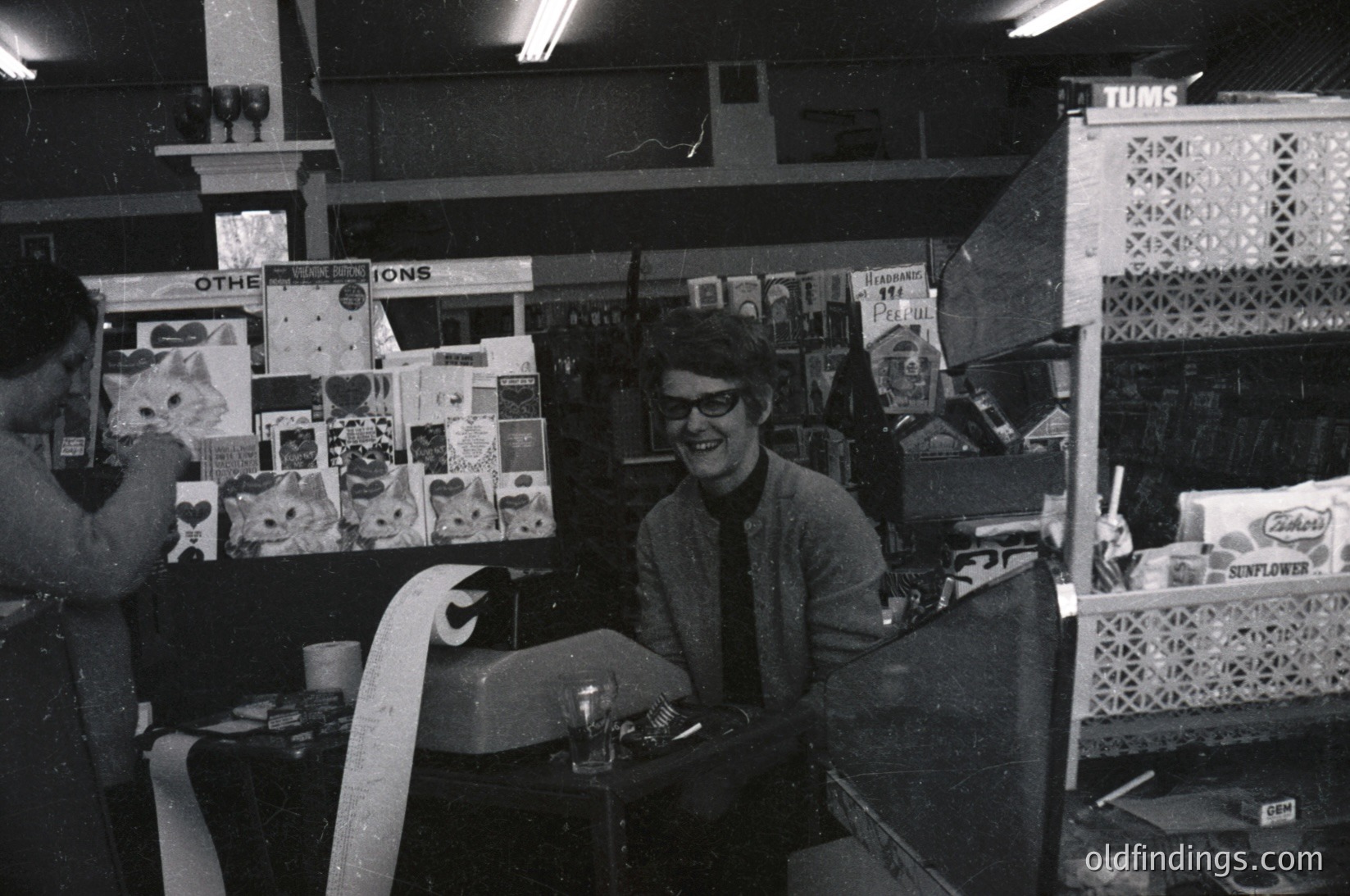 Vintage 1970s-era record store interior with shelves stocked with vinyl albums, posters, and memorabilia. Man in glasses and patterned shirt poses behind counter displaying "OTHELLIONS" album. Shelving includes brands like Tums and Sunflower. Warm lighting and cluttered aesthetic reflect mid-century pop culture.
