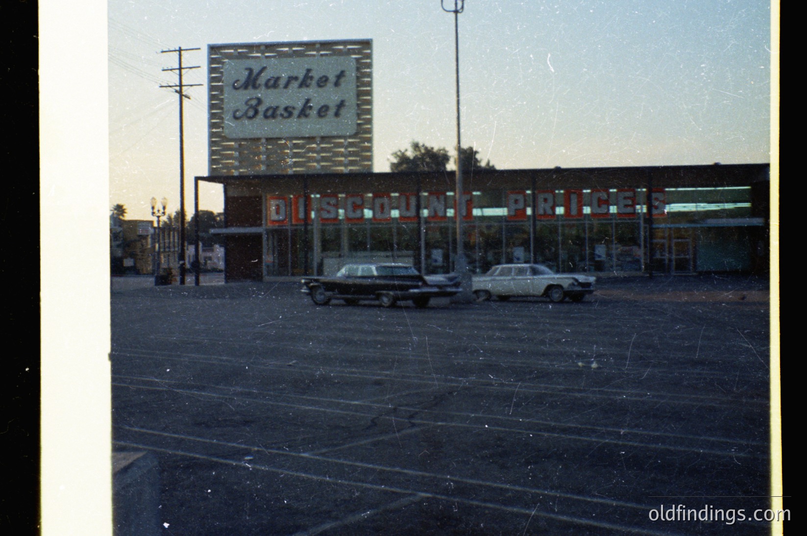 Vintage "Market Basket" discount store signage with retro neon "DISCOUNT" lighting, mid-20th century American suburban setting. Two classic sedans parked in empty lot, framed by telephone poles and low-angle lighting.