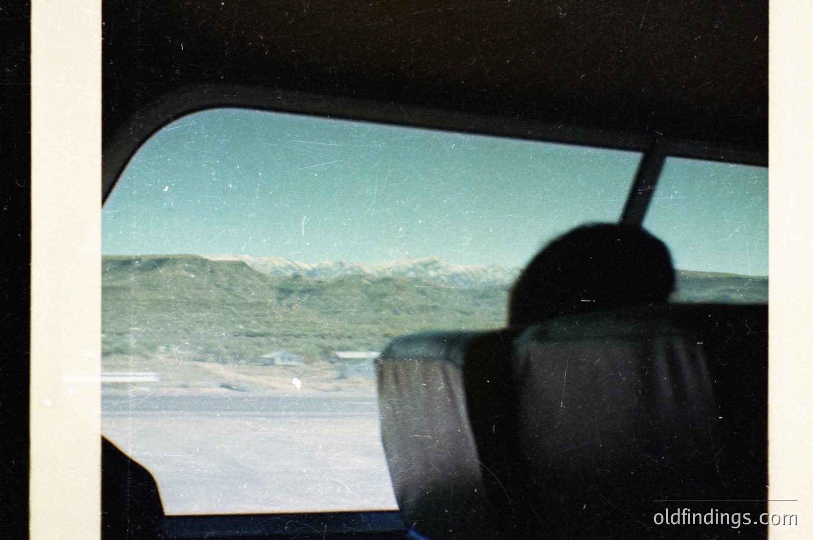 Vintage car windshield view showing snow-capped mountains and a coastal road, likely taken in the 1970s. Distorted lens effect highlights nostalgic travel aesthetic.