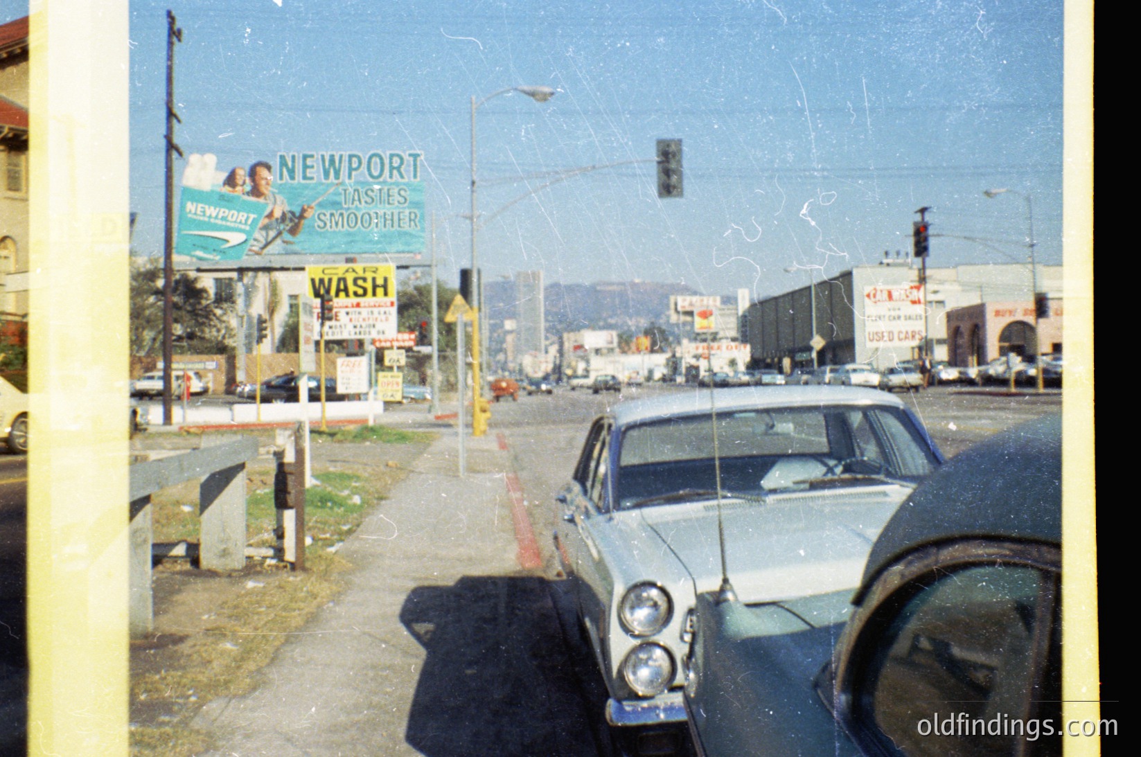 Vintage street scene from mid-20th century, likely 1960s–70s. Classic billboard advertising "Newport Cigarettes" with "Lights Smoother" slogan dominates left side. Mid-century car with dual headlights parked at intersection, surrounded by traffic lights and utility poles. Urban landscape with distant mountains and commercial signage ( )
