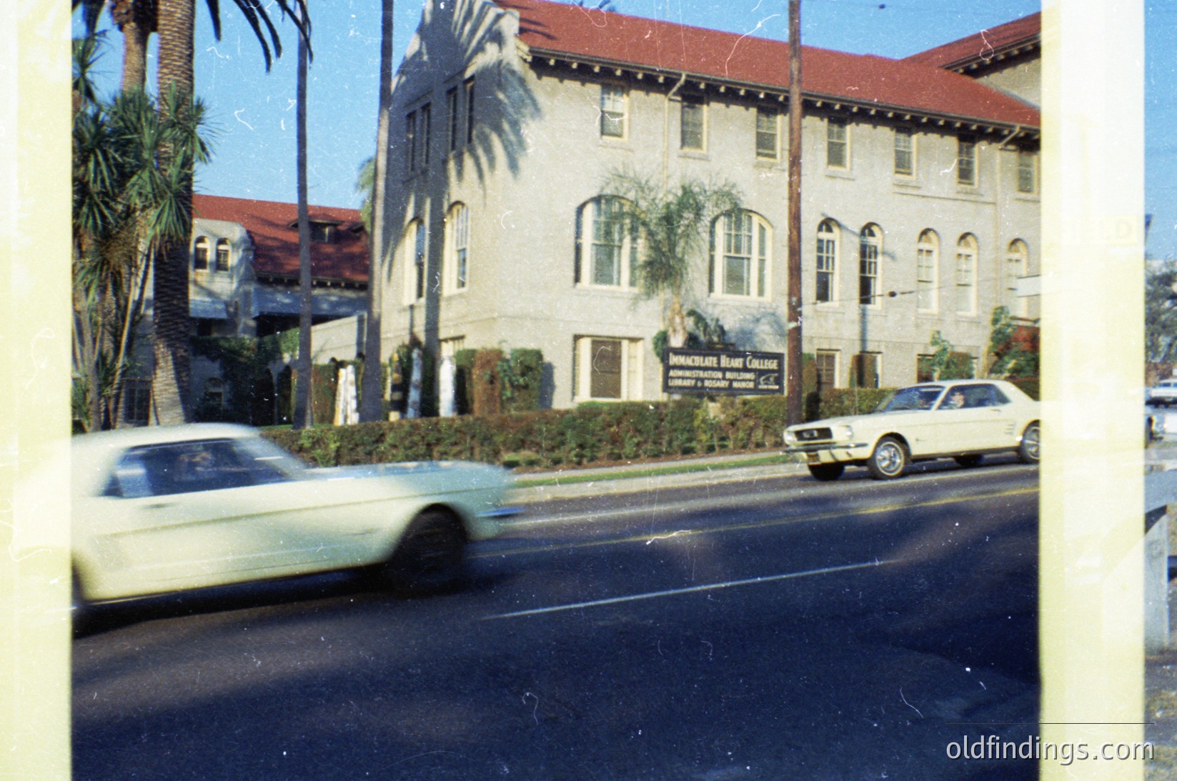 Vintage 1970s-era motel signage ("Sunset Inn Motel") on a two-story stucco building with arched windows, palm trees, and classic American roadside architecture. Blurred vintage car (likely a Ford or Chevrolet) driving past. Warm, nostalgic color tones.