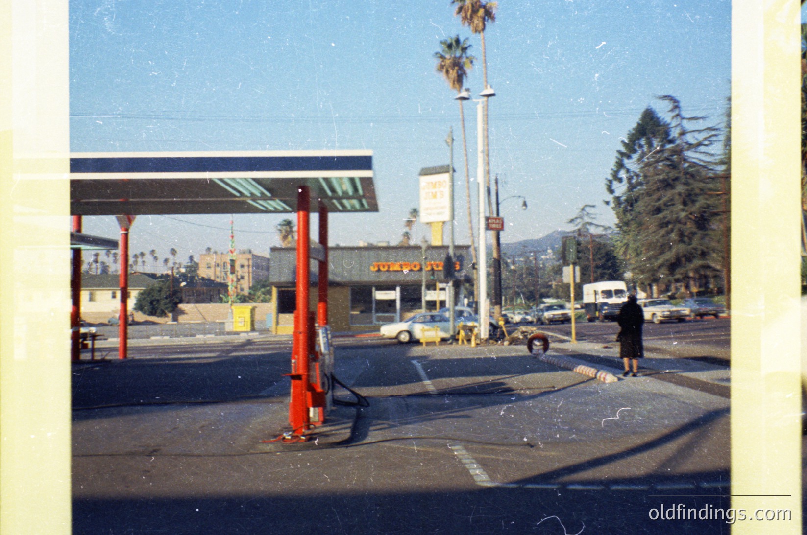 Mid-century gas station with red canopy and white lettering, likely . Palm trees and vintage cars suggest a sunny coastal or suburban setting. Signage includes "76" and "Shell" branding. Single person in dark coat stands near pump, hinting at midday refueling.