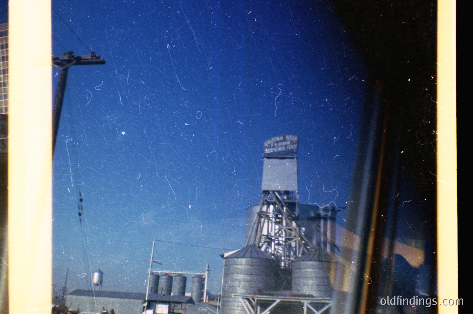 Vintage industrial scene featuring a grain elevator with "A. G. Sperry & Sons Co." signage, likely mid-20th century. Silhouetted against a clear blue sky, with cylindrical storage tanks and utility poles in the background. Film grain and slight blur enhance nostalgic vintage aesthetic.