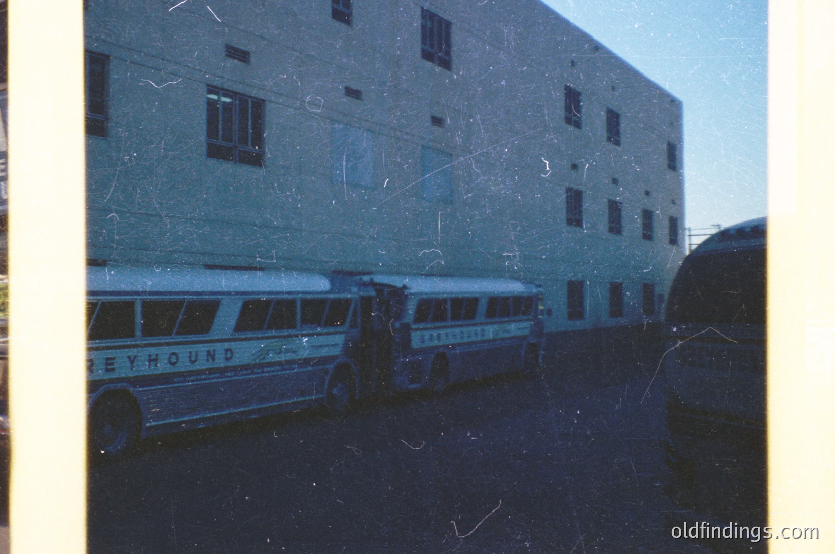 Vintage 1960s-70s **Greyhound bus depot** with two white buses bearing "Greyhound" branding parked in front of a utilitarian concrete building. Aged film grain and sepia tone enhance nostalgic atmosphere.