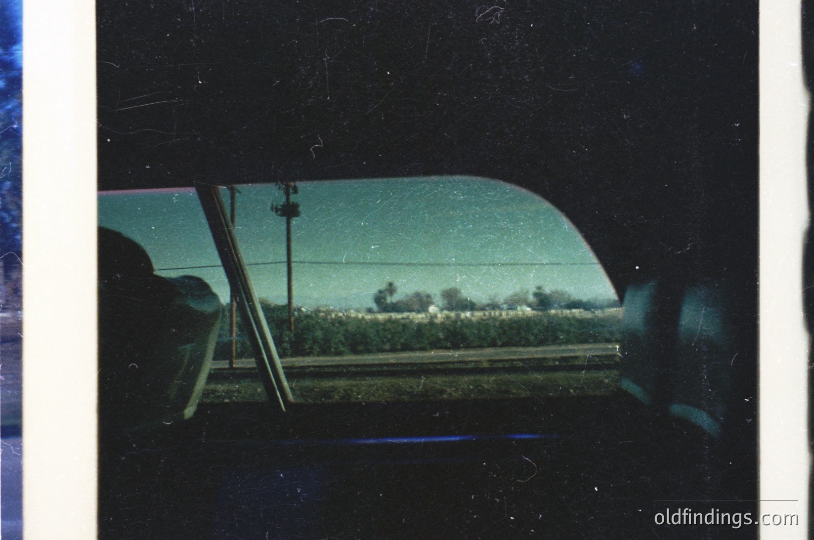 Vintage car interior shot through windshield, showing blurred rural landscape with dense greenery and a faint road. Tinted glass and visible interior trim suggest mid-20th century design.