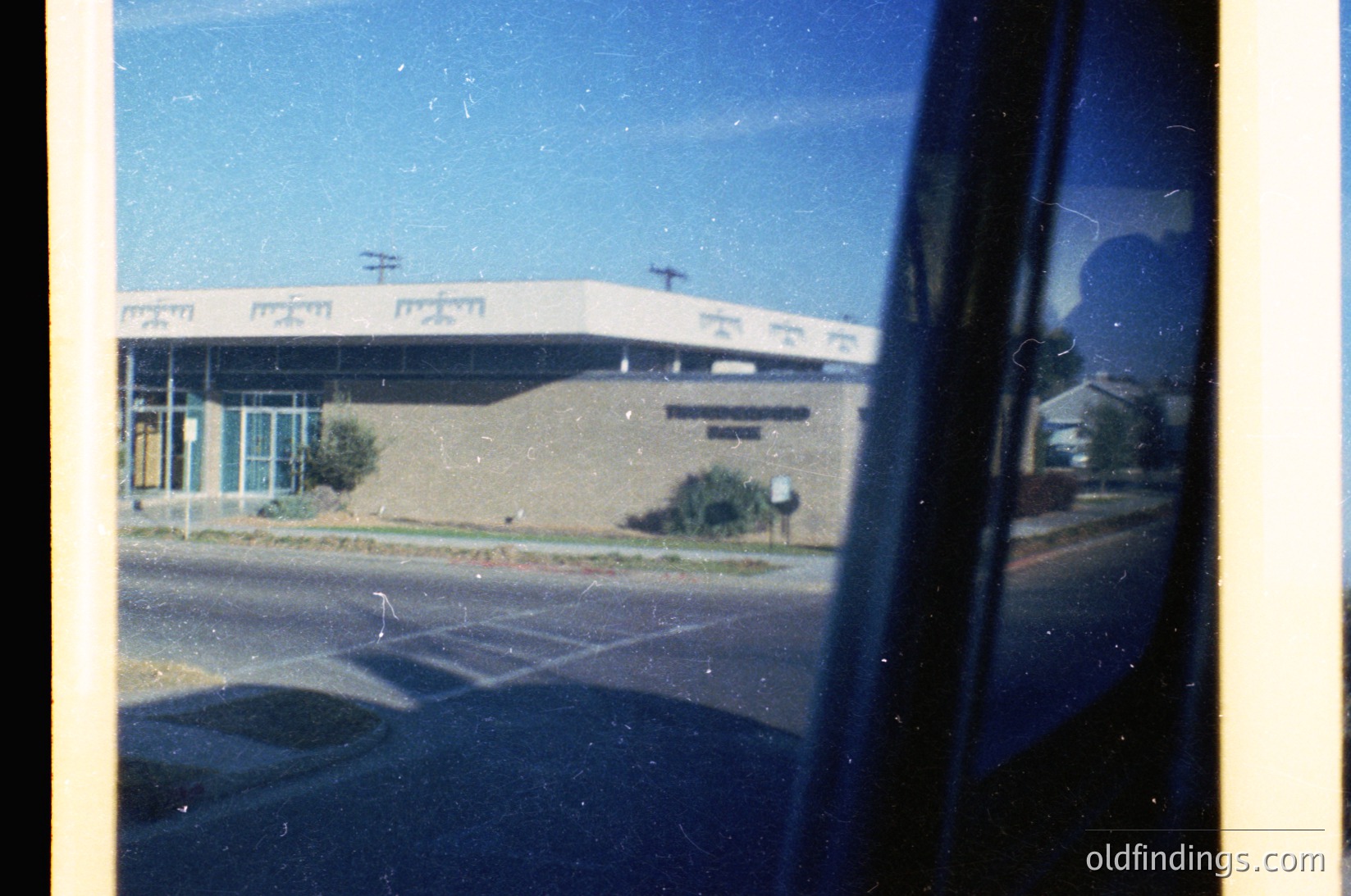 Mid-century modern building with curved concrete roof and large windows, viewed through a vehicle window. Likely a public or institutional structure, possibly a library or community center. Vintage color film grain and reflections add nostalgic texture. Residential area with utility poles in background.