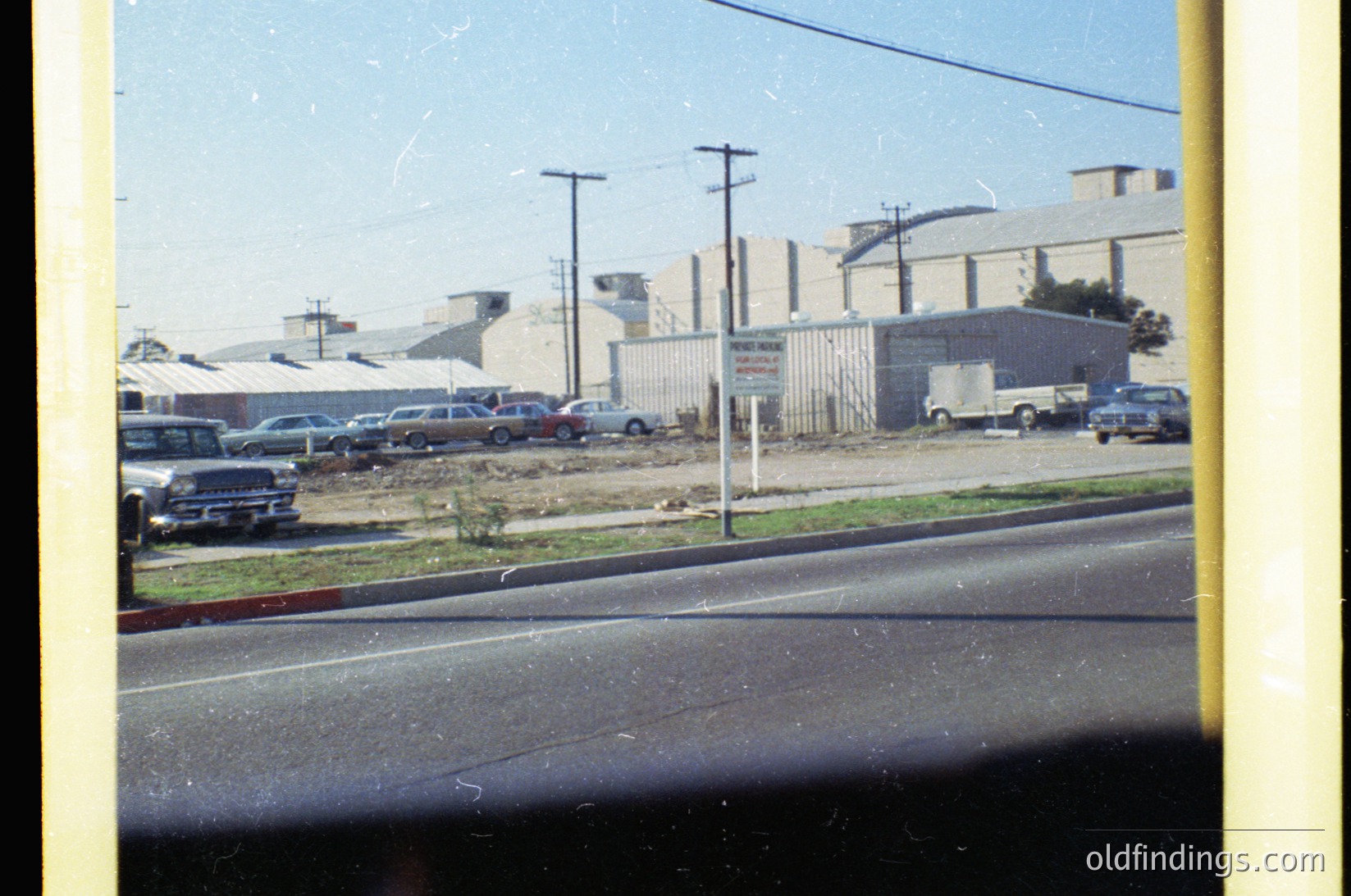 Vintage industrial site with mid-century concrete buildings and parked classic cars (1960s-70s). Overhead wires and utility poles frame the scene. Faded color film captures dusty, arid surroundings.