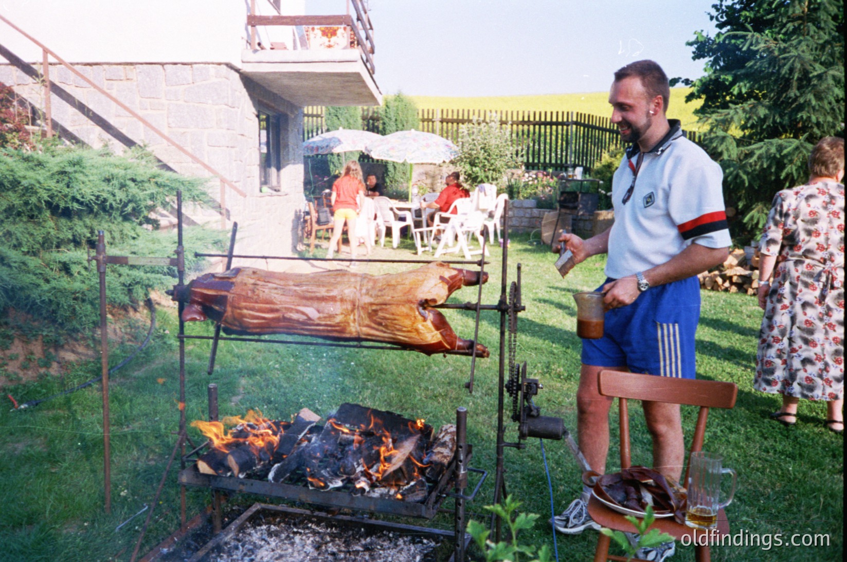 Whole pig roasting over open flame in backyard setting, 1970s-1980s. Man in Adidas track pants and polo shirt tends to fire, while guests relax under umbrella. Concrete block house with balcony in background.
