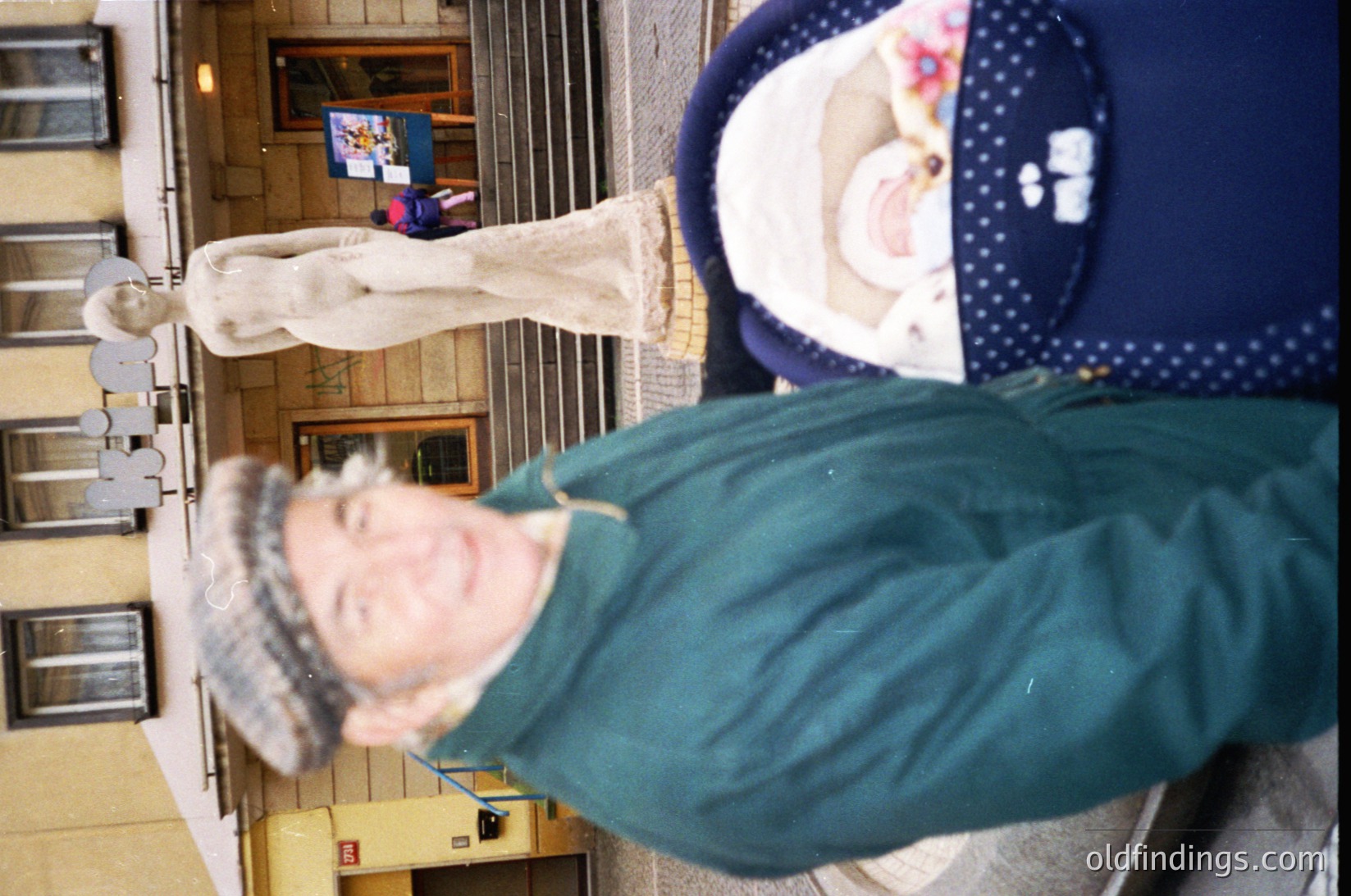 Vintage mirror selfie of two individuals in an indoor hallway, likely a 1980s-1990s setting. The person in foreground wears a teal jacket and white headscarf, while the blurred background figure wears a patterned blouse. Yellow caution tape and a framed poster with indistinct text are visible.