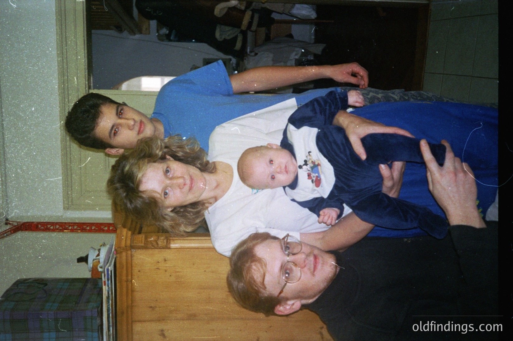 Family portrait in a dimly lit indoor setting, likely a 1990s home. A man in glasses and a dark shirt sits on a wooden bench, holding a baby in a white onesie with a cartoon character. Behind him, a woman in a white top and blue jeans leans forward, while a young man in a blue shirt stands behind her. The background shows tiled walls and a doorway with a red stripe.
