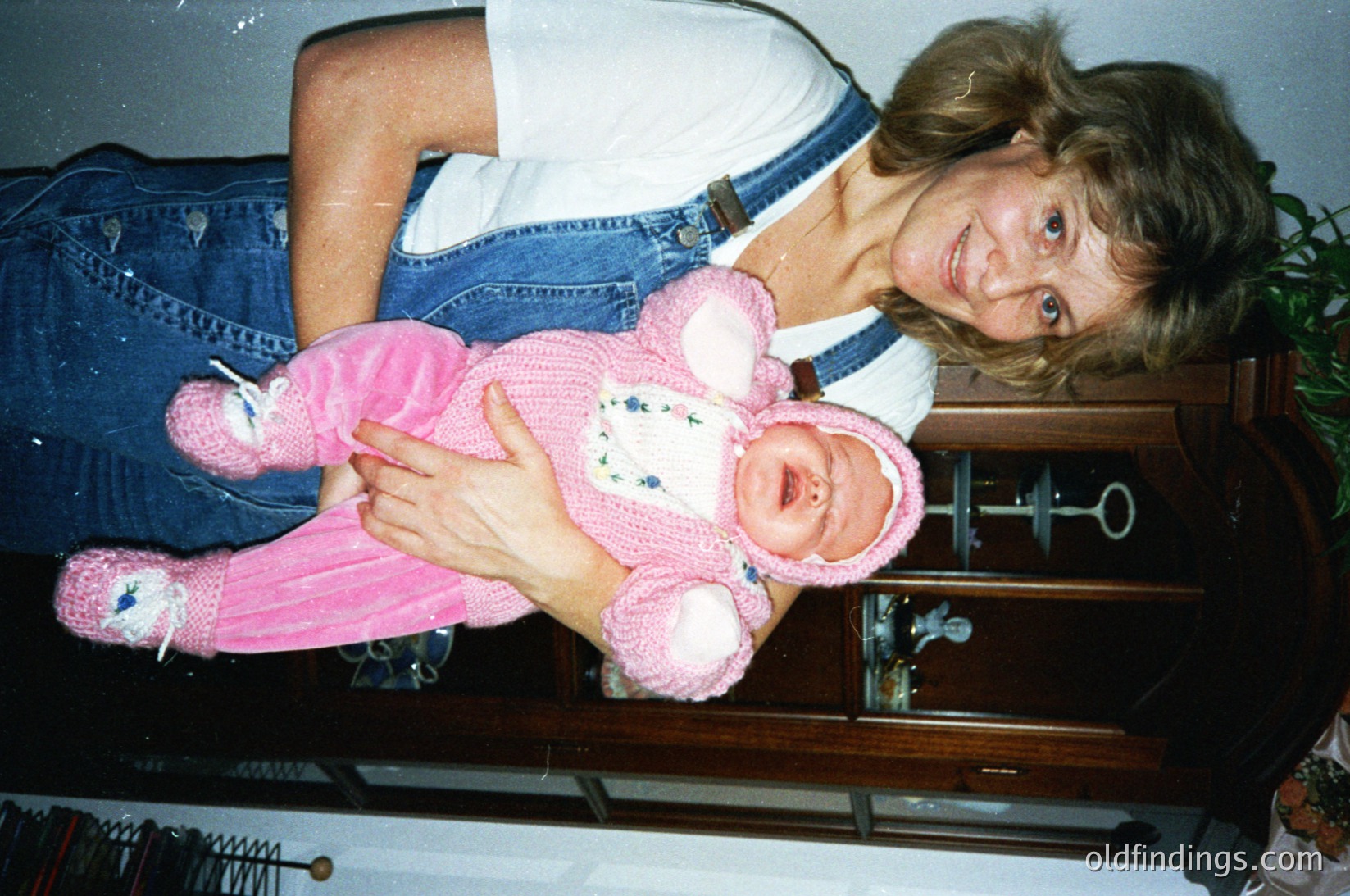 A woman in a denim jacket and blue jeans poses with a baby dressed in a vintage pink knitted outfit, likely from the **1980s–1990s**. The setting appears to be an indoor room with a wooden dresser and potted plant in the background. [Vintage family portrait with baby in 1980s-style pink knitwear ] --- *Note: If this image is for archival purposes, the outfit and hairstyle suggest a Western context, likely North America or Europe.*