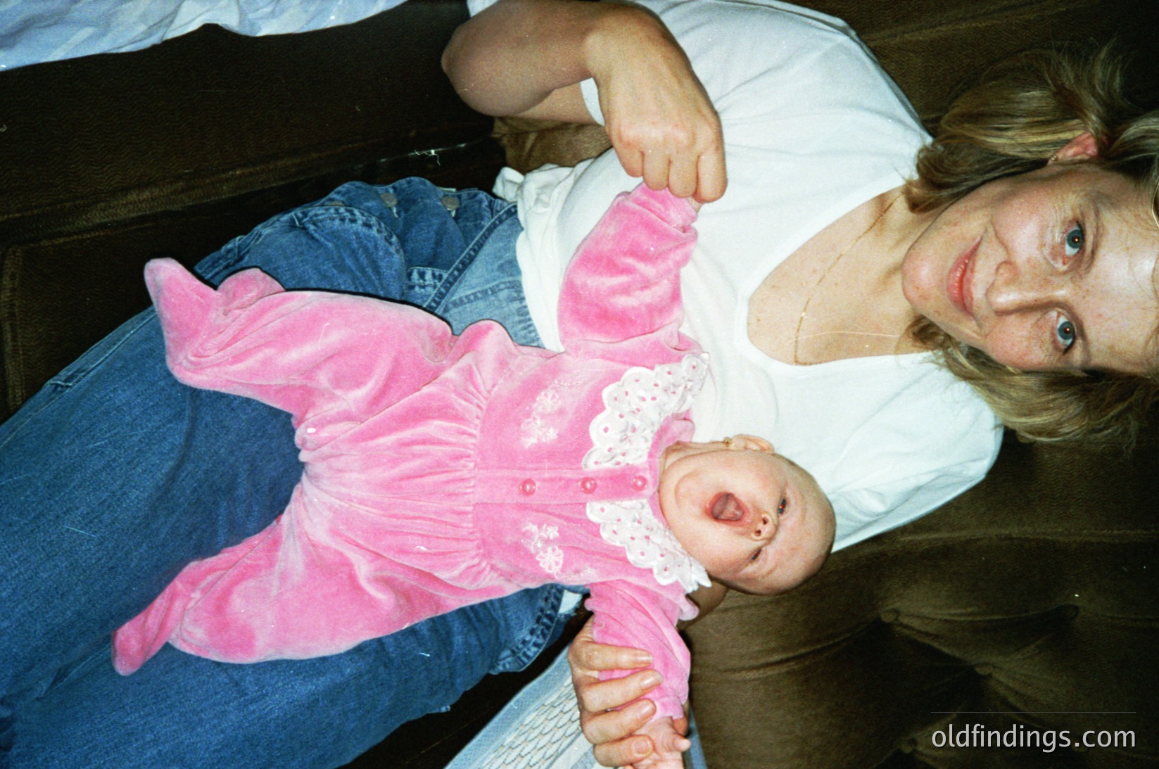 A woman holds an infant dressed in a vintage-style pink lace romper, seated on a wooden bench. The scene suggests a casual, indoor family moment, likely from the 1990s–2000s.