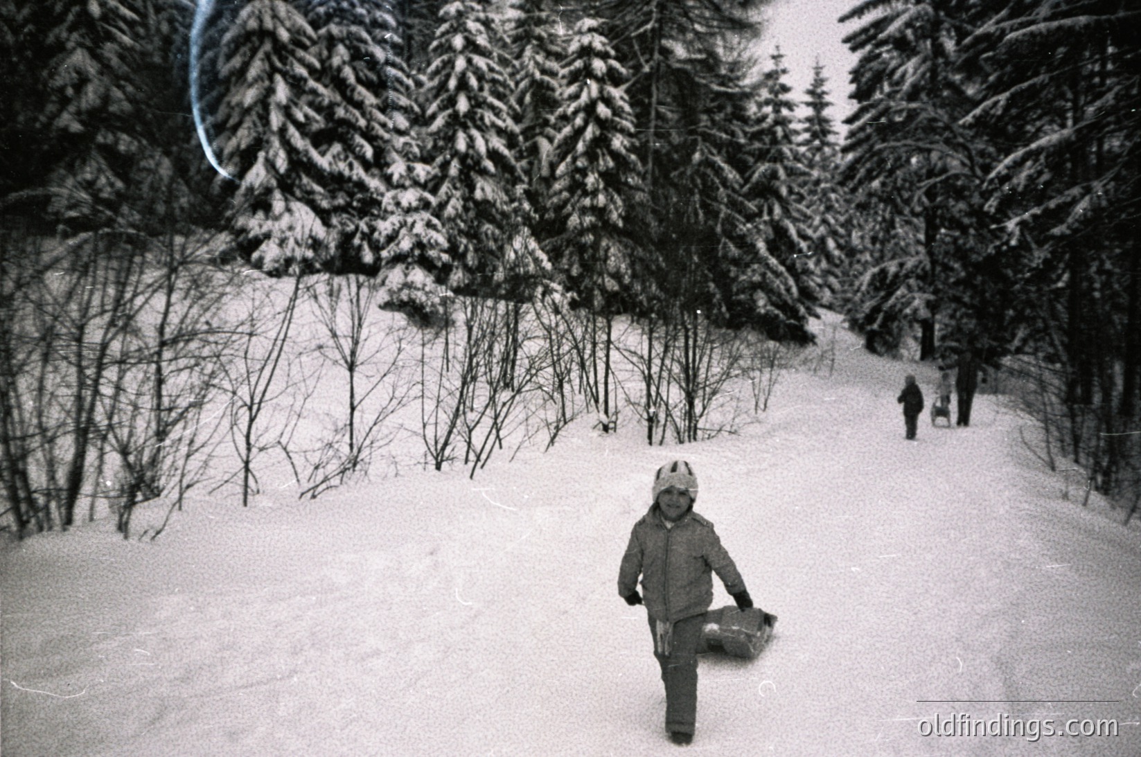 Black-and-white snapshot of a child skiing on a groomed trail surrounded by dense coniferous forest. Snow-covered slopes and distant skiers visible. Likely mid-20th century alpine resort.