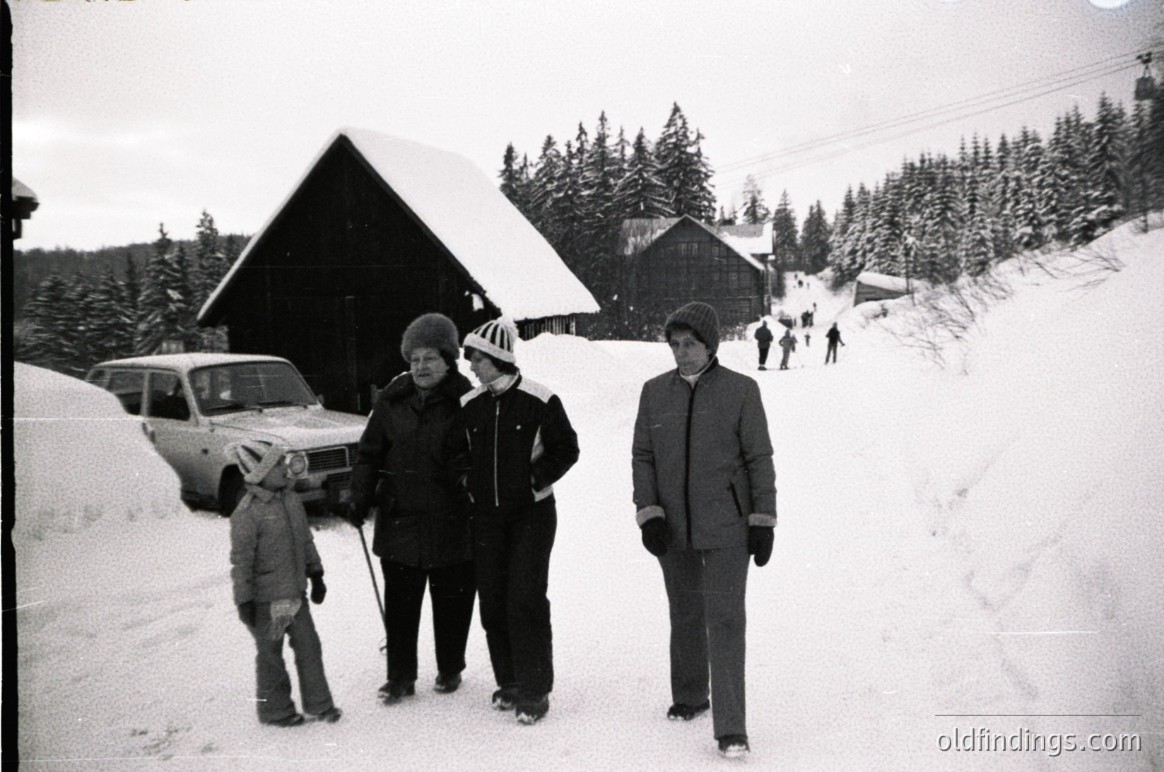 Black-and-white snapshot of a winter outing in a snowy alpine village, mid-20th century. Three adults and a child pose in winter attire—wool coats, hats, and gloves—near a vintage car parked on snow-covered ground. Snow-laden pine trees and rustic wooden chalets frame the scene.
