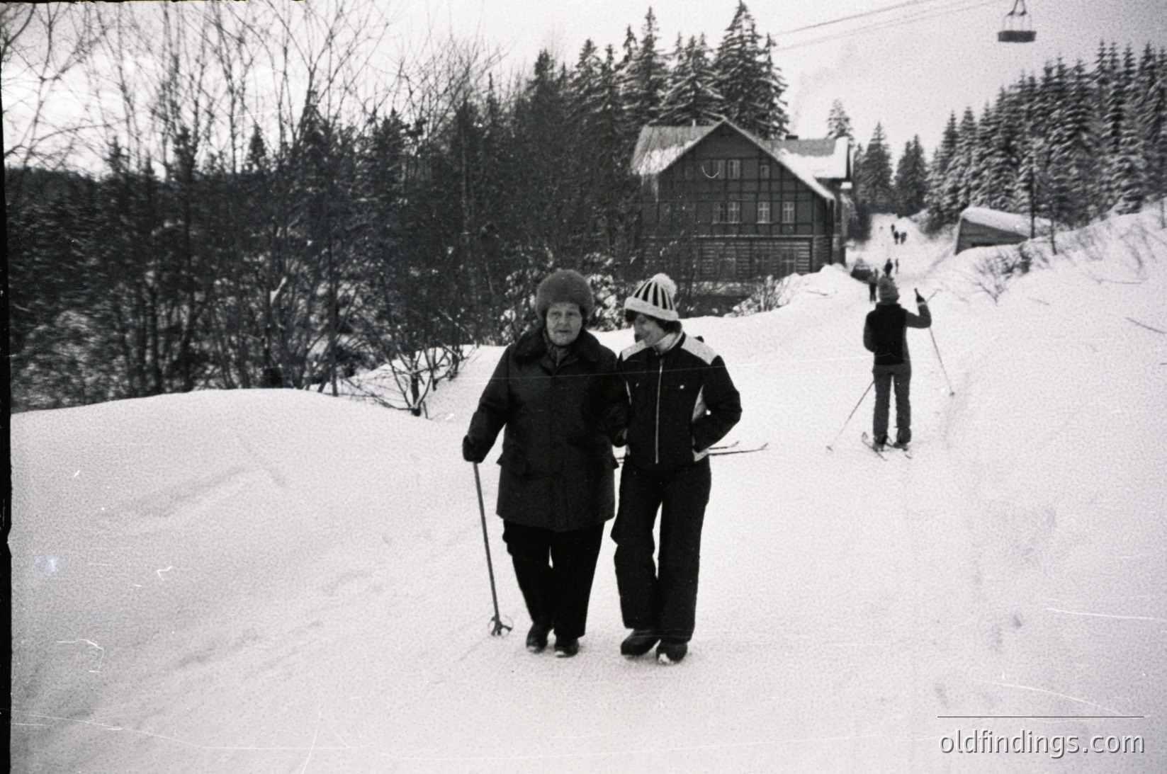 Black-and-white ski resort scene from mid-20th century, likely 1950s–1960s. Two adults in vintage winter gear—heavy coats, hats, and ski poles—walk along a groomed slope beside a wooden chalet-style building. Snow-covered forest and ski lift cables in background.