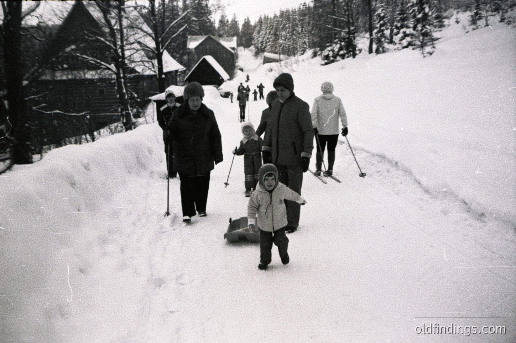 Family skiing on groomed trail in snowy alpine village, mid-20th century. Adults in vintage winter gear (coats, hats) guide child on sled. Wooden chalets and evergreen trees frame the scene. Likely European,