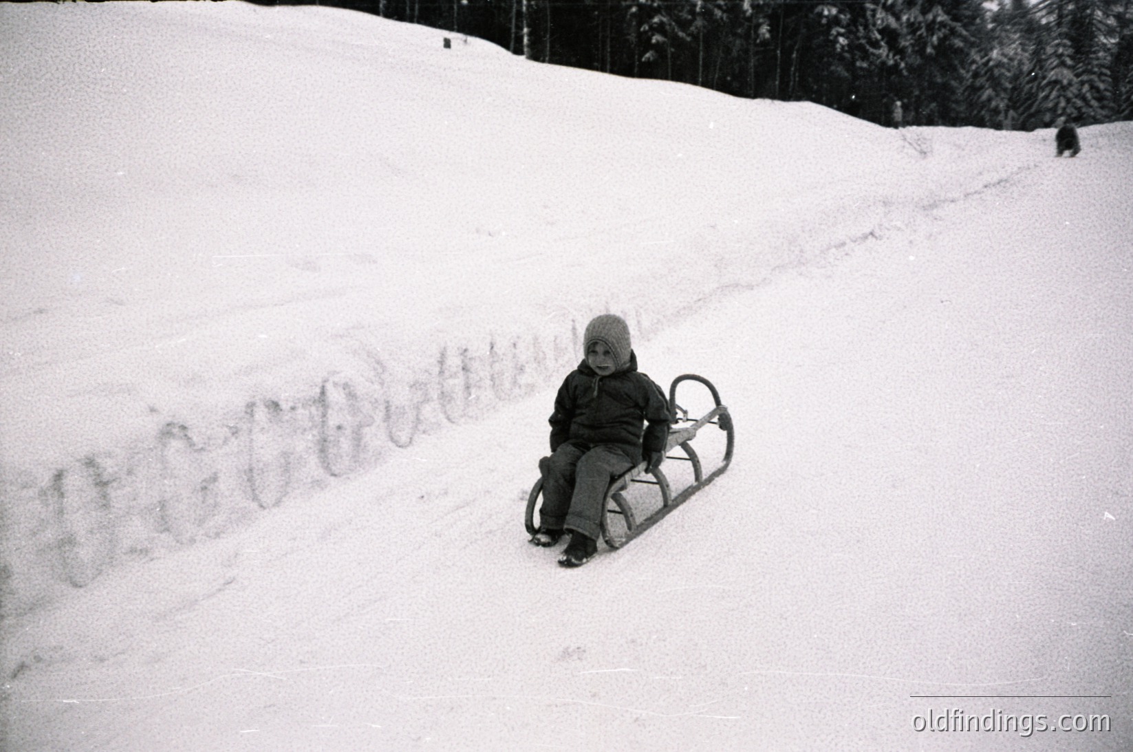 Child in vintage-style sled on snow-covered slope, mid-20th century winter scene. Snow tracks and evergreen trees frame the composition. Classic black-and-white winter play