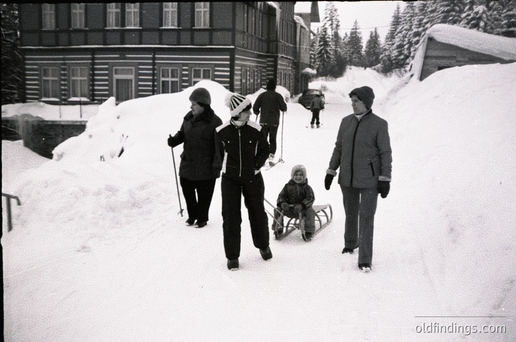 Four adults and a child in winter attire navigate snow-covered alpine terrain, likely a ski resort. Wooden chalet-style buildings with white-framed windows line the background. The child rides a sled, while others carry winter gear. Mid-20th century (1950s–1970s) alpine lifestyle captured.