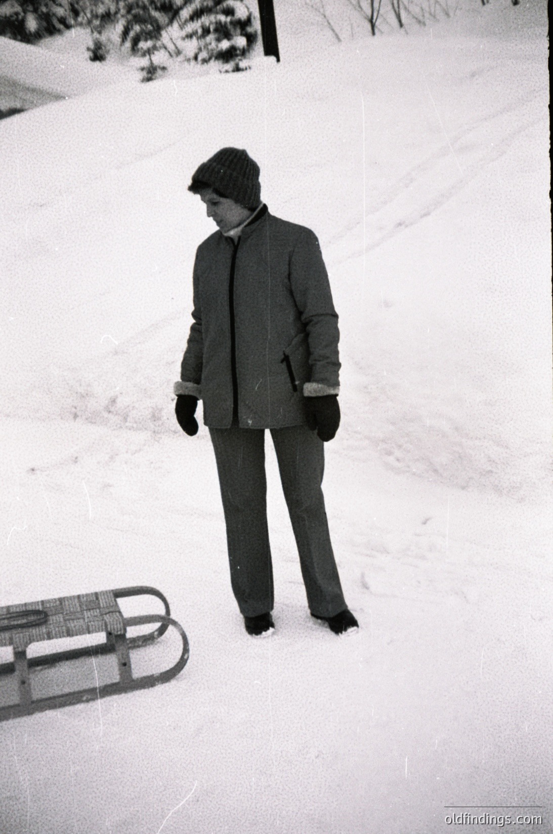 Black-and-white winter scene featuring a child in mid-20th century winter attire—knit cap, padded coat, and trousers—standing on snow-covered ground beside a wooden sled. Snow blankets the ground, and bare trees frame the background. Likely or winter play setting.