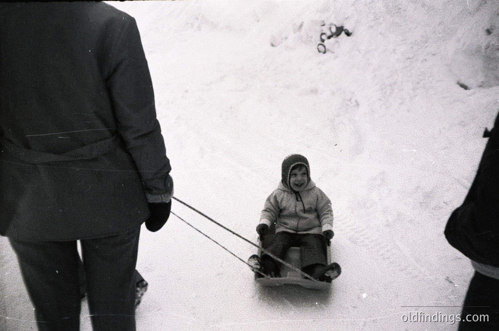 A child in a makeshift sled (rope-wrapped log) is pulled by an adult in mid-winter, likely 1960s–1980s. Snow-covered ground and partial winter coats suggest cold-weather recreation.