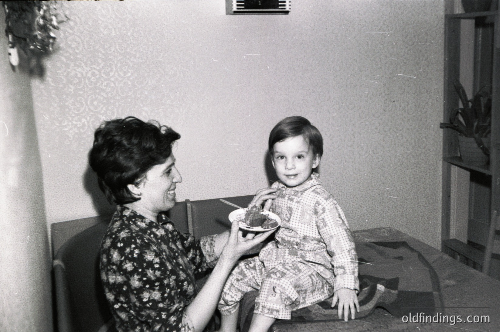 A mid-20th century indoor moment: an adult feeds a young child a small bowl of food in a modestly furnished room. The woman wears a floral-patterned blouse, while the child sits on a patterned rug. Shelves with decorative items and a chandelier-style light fixture frame the scene. Likely 1950s–1960s domestic life.