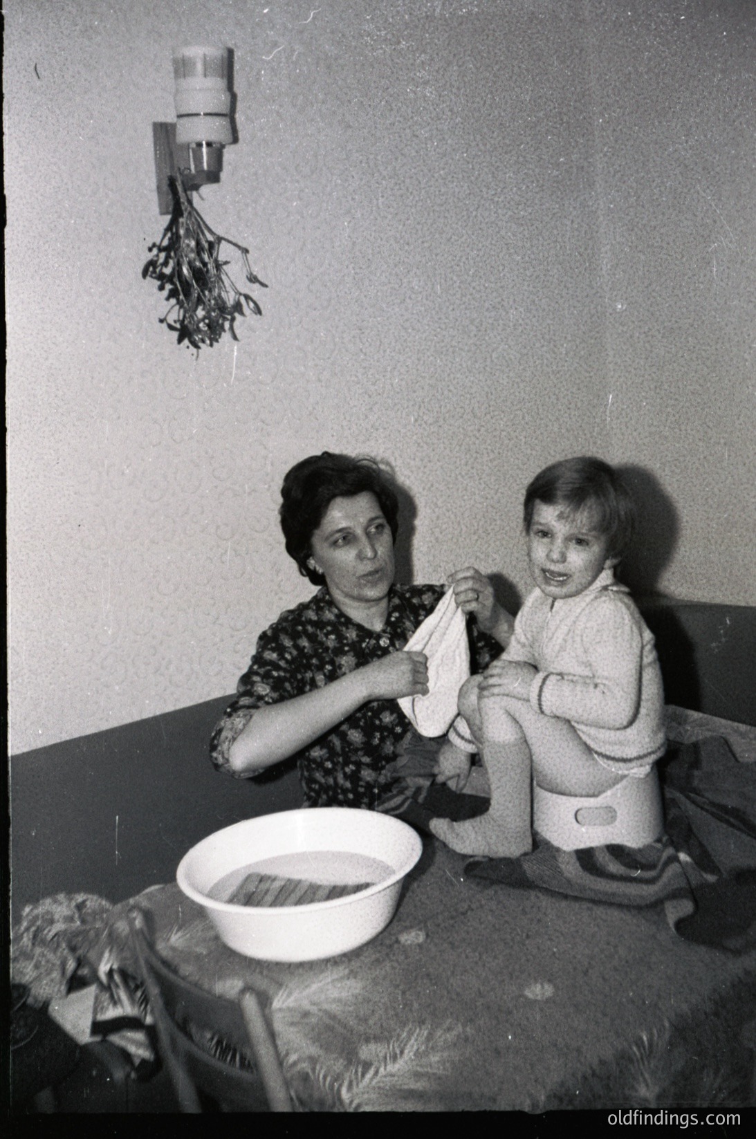 A black-and-white photo capturing a mid-20th-century domestic moment: a woman in patterned dress assists a toddler in a training potty. Decorative pine branch wall hanging and a bowl of laundry on a table suggest a modest, homey setting.