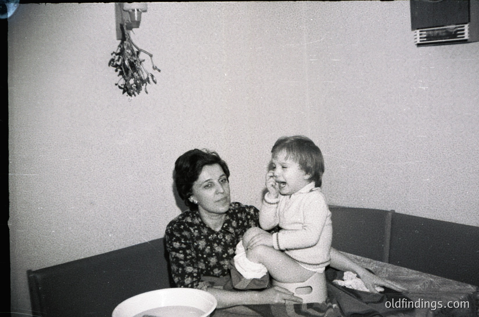 Vintage black-and-white photo of a woman cradling a toddler in a modest indoor setting, likely mid-20th century. Patterned floral dress and simple wooden furniture suggest domestic warmth. Decorative dried herbs hung above, hinting at traditional home remedies.