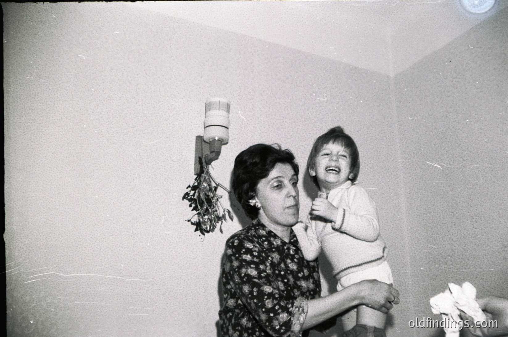 Vintage black-and-white photo of an adult holding a laughing child indoors, likely mid-20th century. Floral wallpaper and a wall-mounted flower holder with dried blooms visible. Simple, intimate domestic scene.