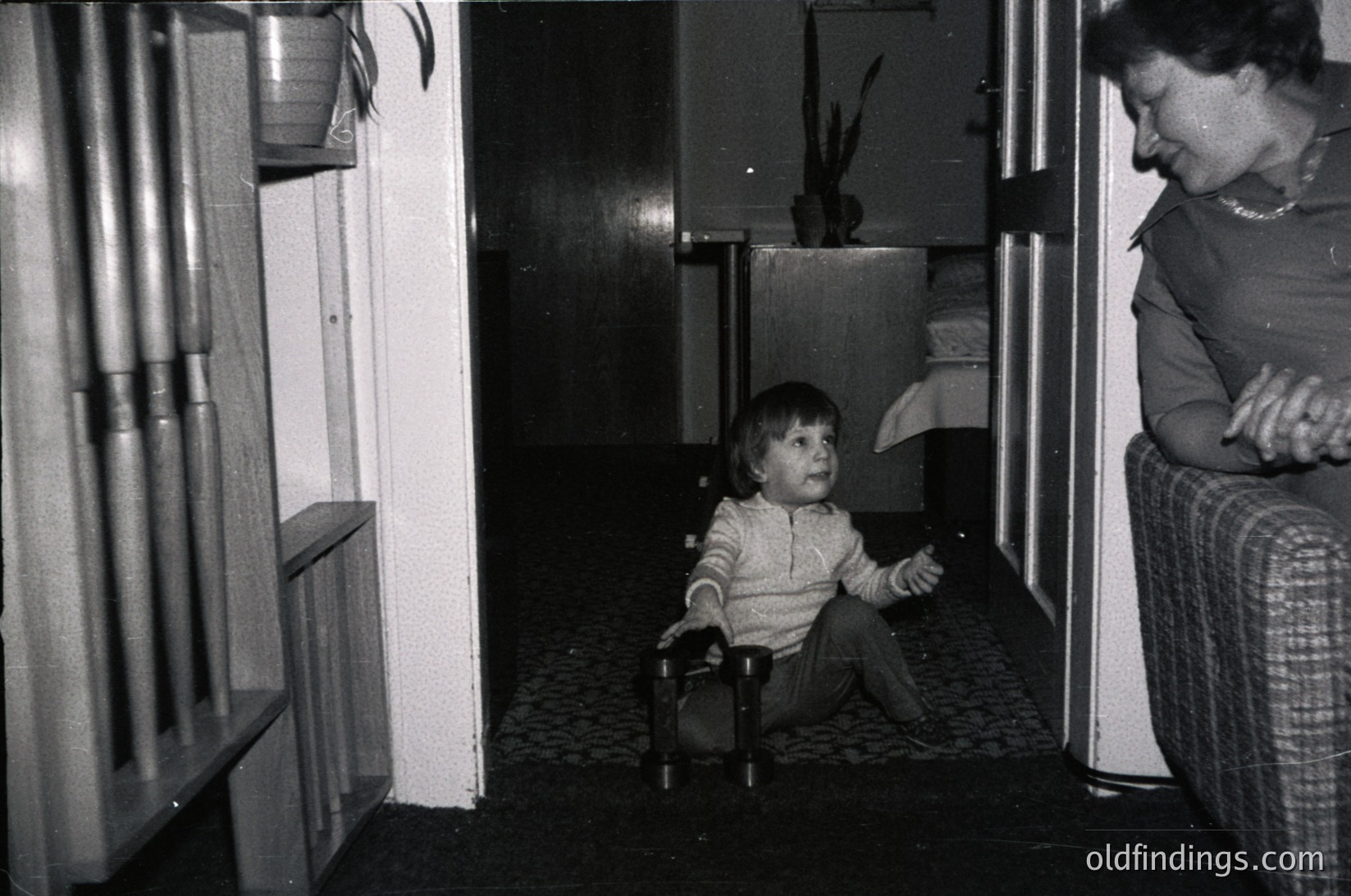 A mid-20th century indoor scene captures a child seated on a highchair in a dimly lit hallway, flanked by a woman in patterned pants. The highchair’s metal frame and wooden seat reflect 1960s-70s design. Adjacent shelves hold cylindrical objects, possibly storage or decorative items. The tiled floor and white-painted walls suggest a residential setting, likely Eastern European based on architectural style. [Vintage highchair in hallway ]