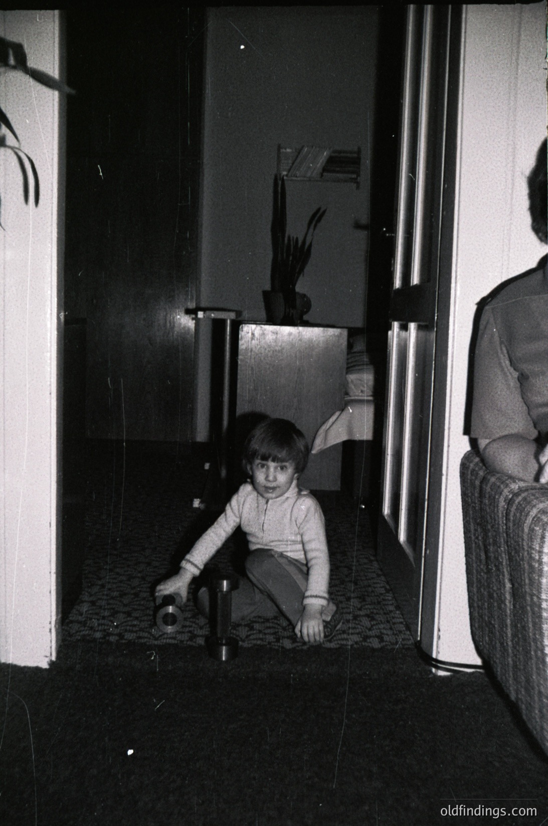 Black-and-white interior shot of a child playing with a small object on tiled floor, framed by white-painted walls and doors. Mid-century domestic setting with minimalist decor—notice the potted plant and stacked bookshelf in background. Likely late 20th century (1960s–1980s).