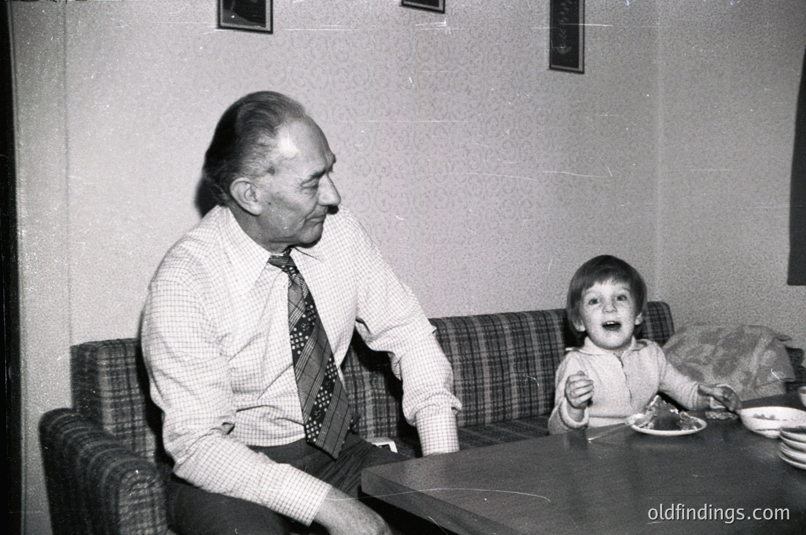 Mid-20th century indoor portrait: elderly man in checkered shirt and patterned tie sits beside a young boy at a wooden table. Boy gestures excitedly toward food, likely 1960s–1970s home or café setting. Warm, candid family moment.