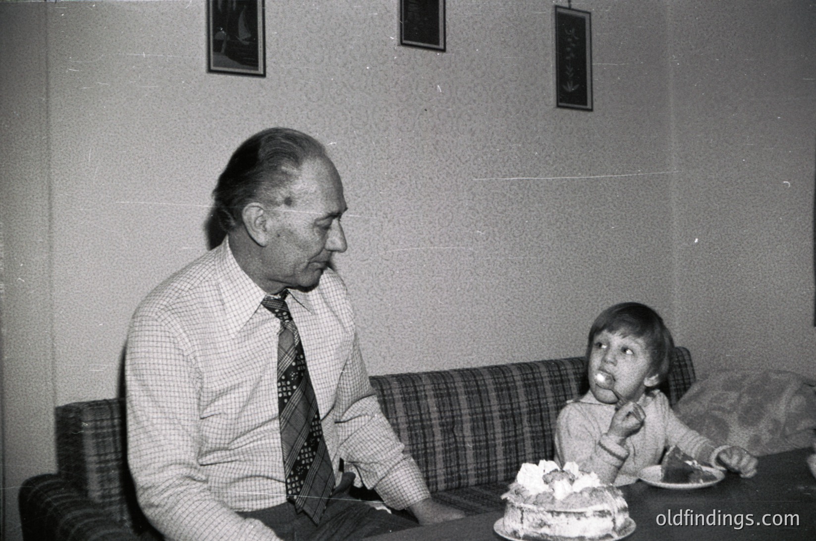 Mid-20th century indoor portrait: an older man in a checkered shirt and tie sits beside a young boy on a plaid sofa, both facing a birthday cake on a table. Plain wall with framed art. Likely 1960s–1970s Western household.