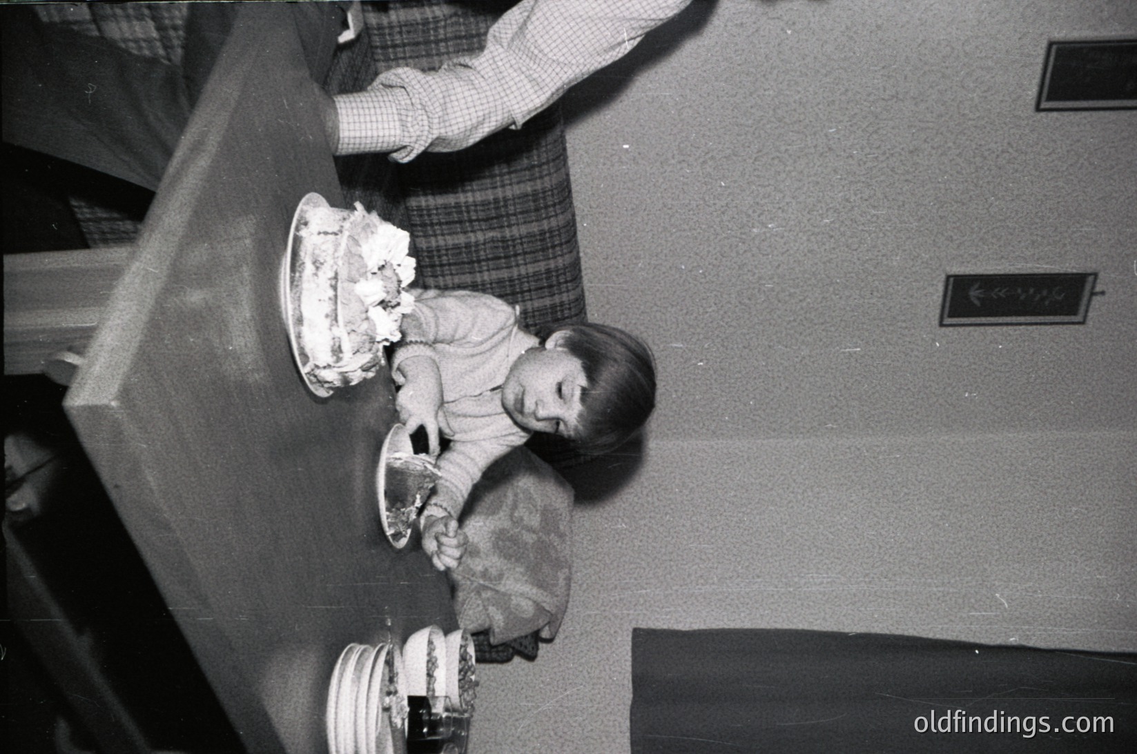 A child in a sweater and skirt sits on a carpeted floor, holding a small cake with lit candles. An adult’s legs in checkered pants appear in the background, suggesting a domestic indoor setting. Mid-20th century (likely 1950s–1960s) based on clothing and lighting. [Child’s birthday celebration in mid-20th century home, vintage ]
