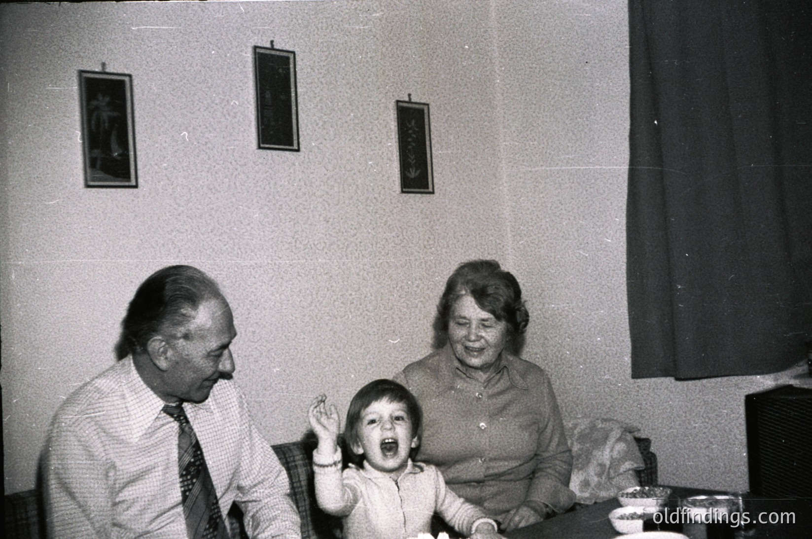 Black-and-white indoor portrait of a family: elderly man in suit, woman in patterned blouse, and child laughing mid-table. Framed religious icons on neutral-toned wall. Mid-20th century domestic setting, likely Eastern European.