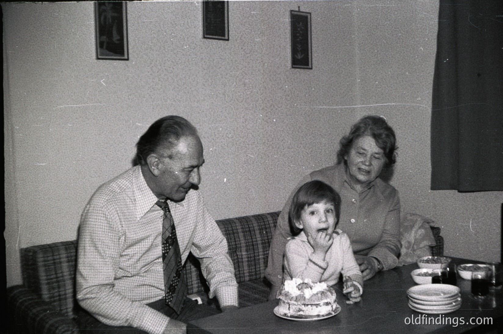Vintage black-and-white indoor family moment: elderly man in checkered shirt and tie, child in white dress eating birthday cake, seated woman in dark sweater. Plain walls, patterned upholstery, simple tableware. Likely mid-20th century domestic setting.