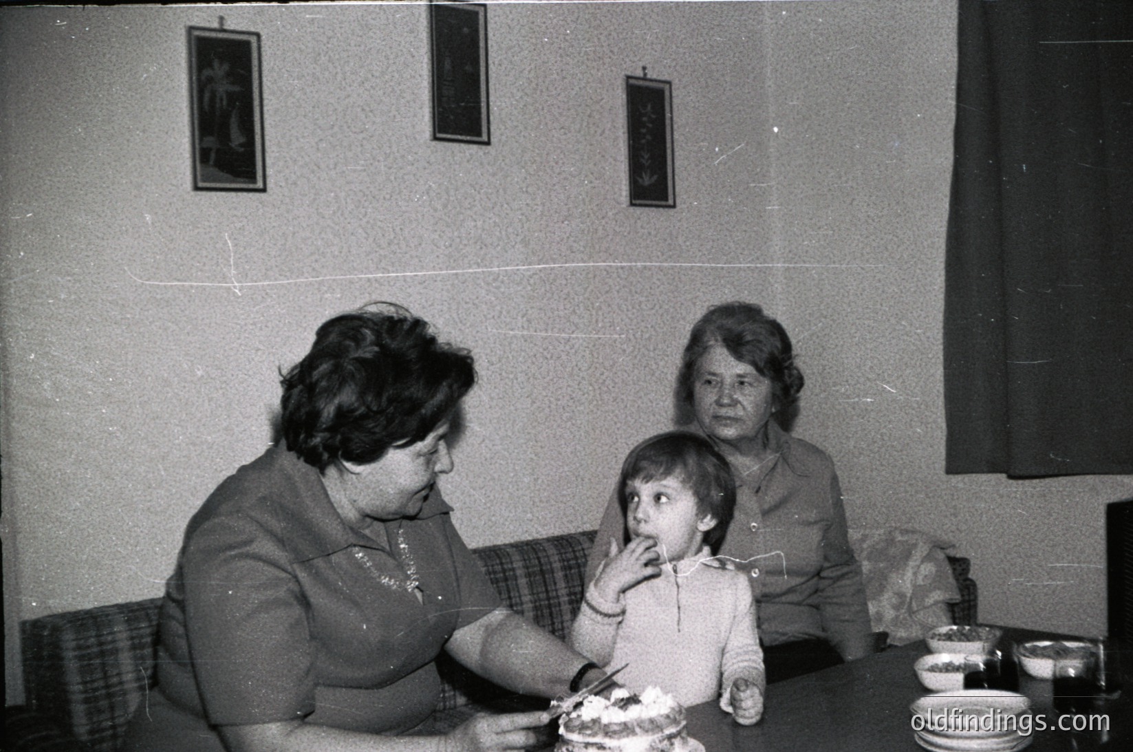 Two women and a young child seated indoors at a table, likely mid-20th century. The woman on the left feeds the child a slice of cake, while the seated woman on the right observes. Plain wall with framed religious icons, simple wooden table with stacked plates.