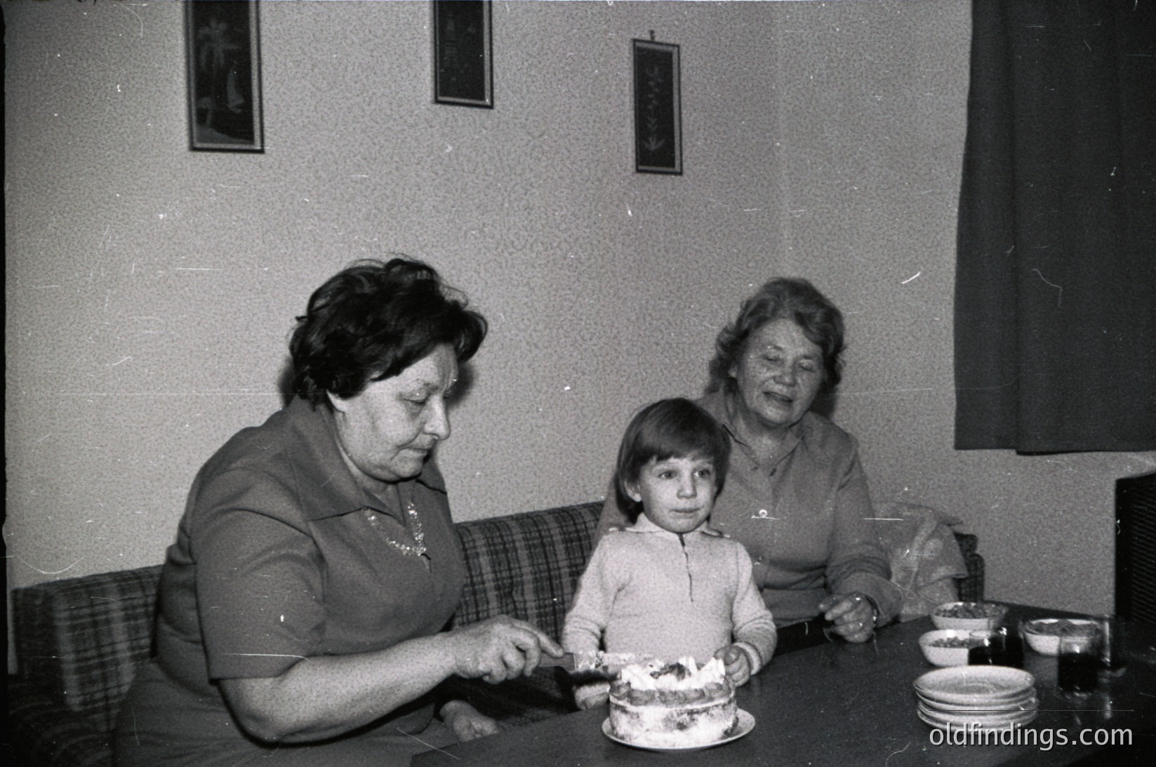 Black-and-white indoor scene featuring three generations: two women (likely mother and grandmother) and a child at a round table. The woman on left cuts cake with a knife, while the seated grandmother holds a fork. The child stands between them, holding a plate. Plain wall, floral-patterned couch, and simple tableware suggest mid-20th century domestic setting.